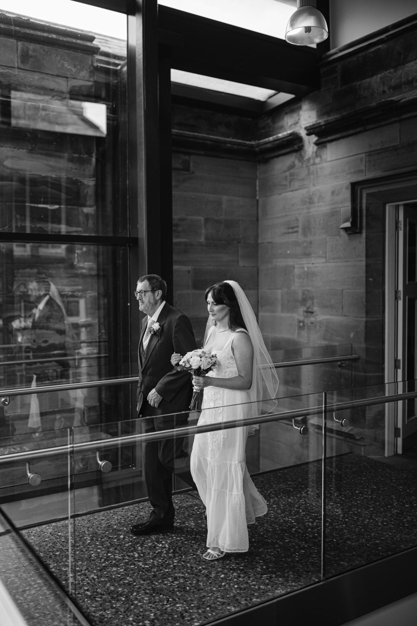 A bride in a white dress and veil holds a bouquet while walking beside an older man in a suit through a modern glass corridor of Carlisle Registry Office.