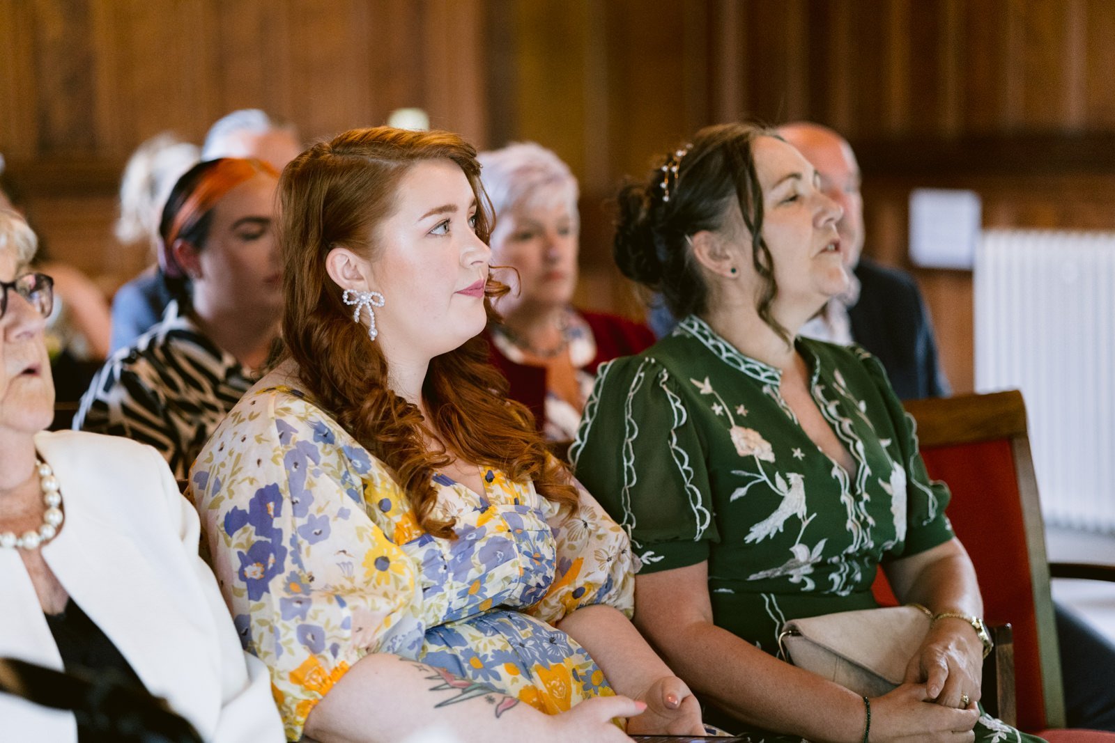 People seated indoors, some looking forward attentively. At the Carlisle Registry Office, a young woman in a yellow floral dress and a woman in a green dress are focused on something out of the frame. Wood-paneled background.