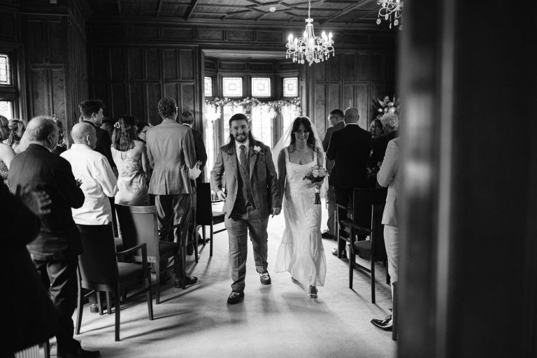 A bride and groom walk down the aisle, hand in hand, in a wooden-paneled room filled with seated guests following their Carlisle Registry Office wedding ceremony.