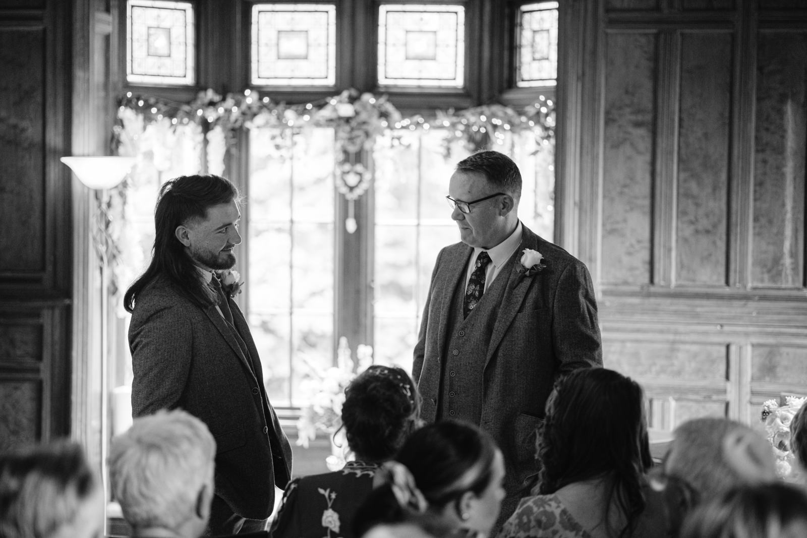 Two men in suits stand facing each other while conversing indoors, likely discussing the upcoming wedding. Seated guests are present in the foreground. The room, reminiscent of Carlisle Cathedral, features tall stained glass windows and wooden paneling.