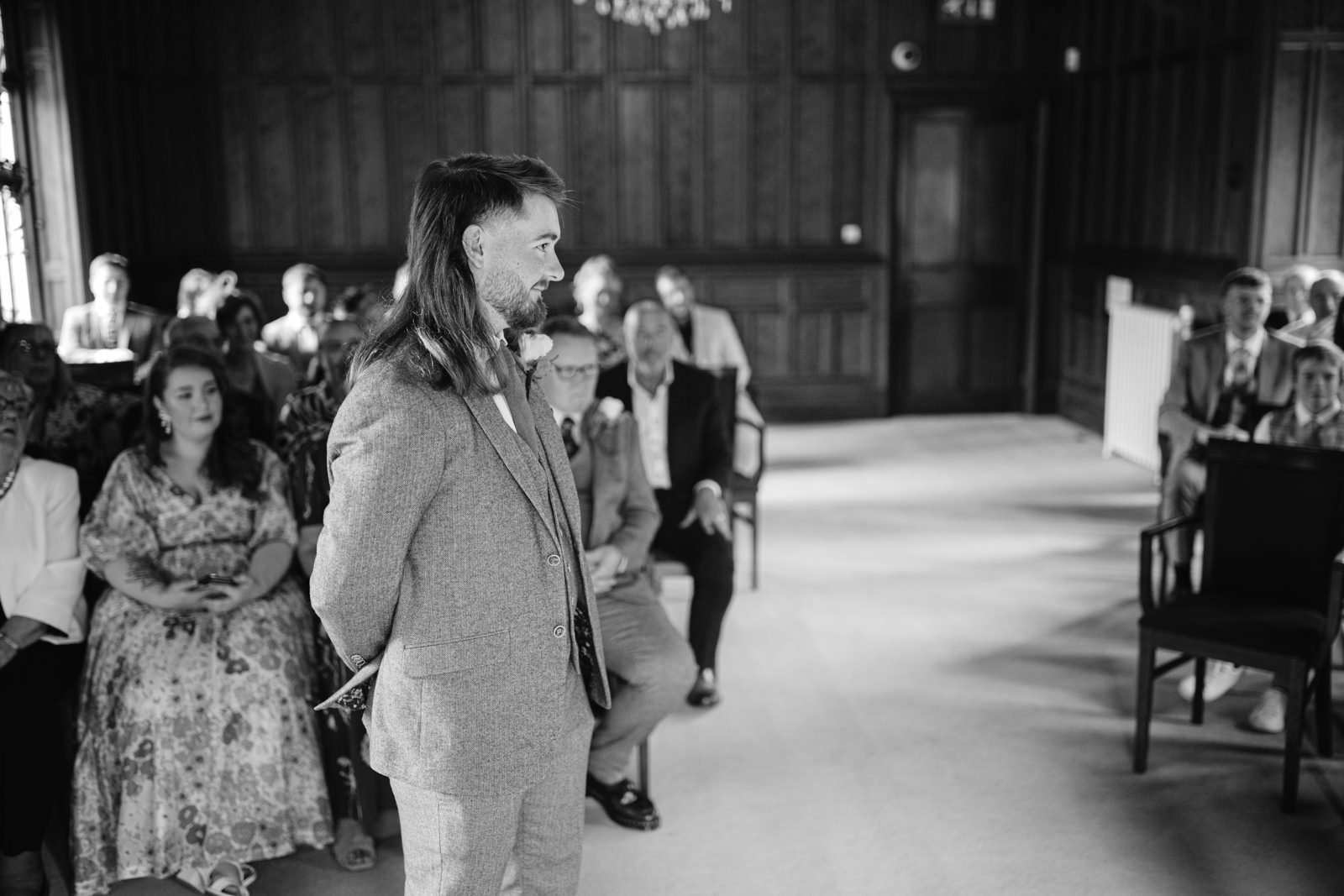 A person in a suit stands in the aisle of a wood-paneled room, addressing a seated audience, much like the serene ambiance found at the historic Carlisle Registry Office.