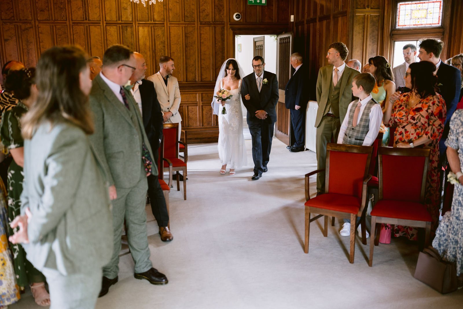 A bride walks down the aisle with an older man in a wood-paneled room at Carlisle Registry Office as guests look on.