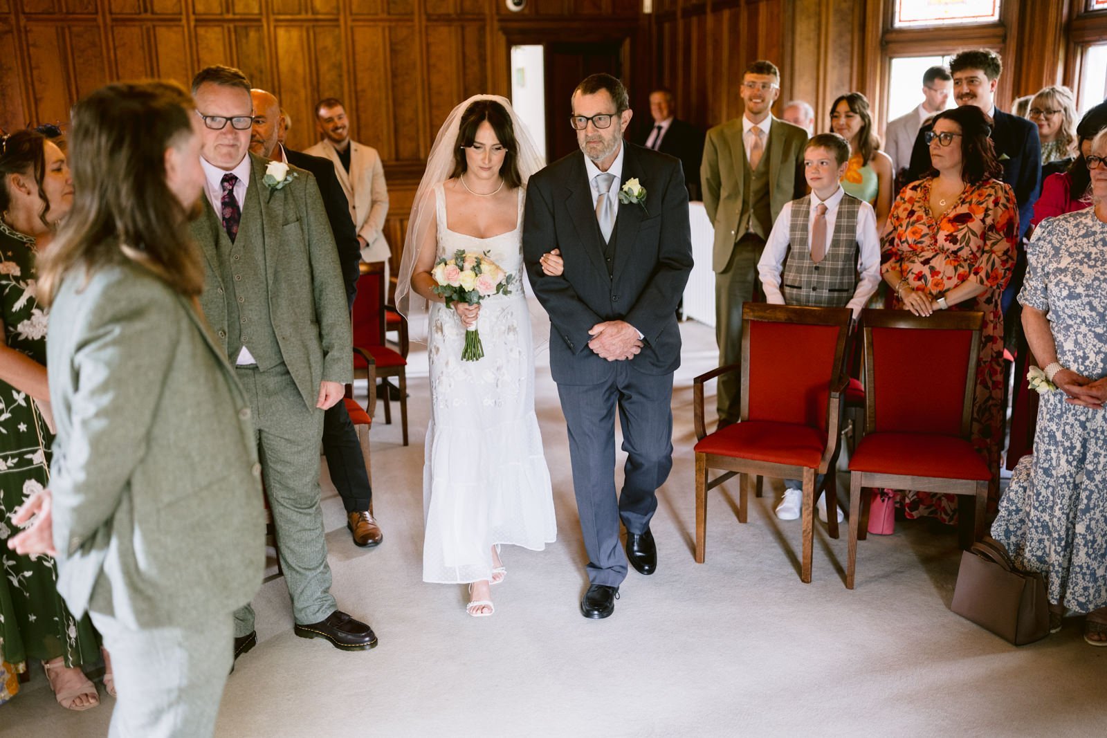 A bride in a white dress, holding a bouquet, walks down the aisle arm-in-arm with an older man in a suit at Carlisle Cathedral, surrounded by standing attendees in formal attire.