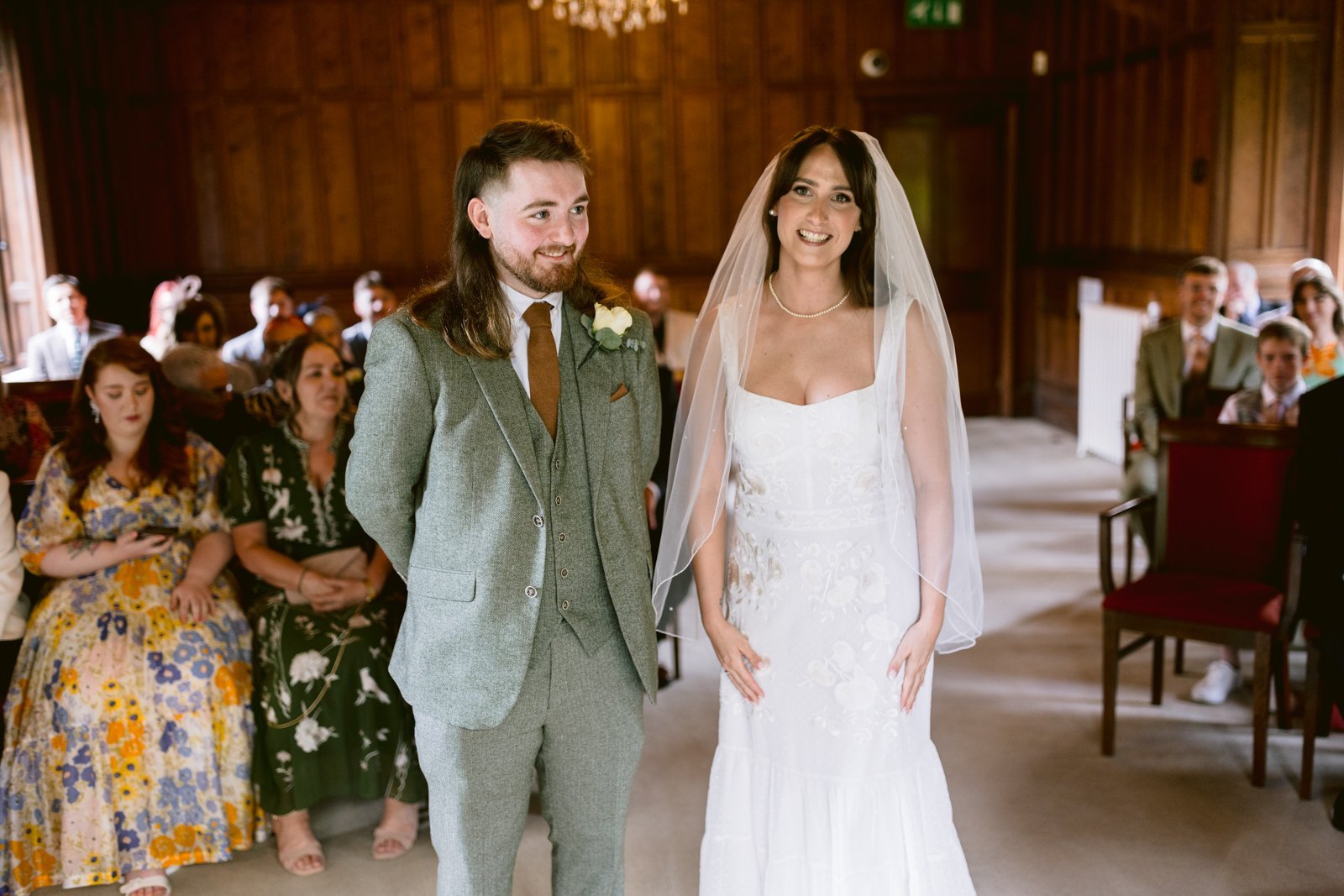 A couple stands side by side, smiling, during their wedding ceremony at the Carlisle Registry Office, in a wood-paneled room with guests seated behind them. The woman is in a white dress and veil, and the man is in a gray suit.