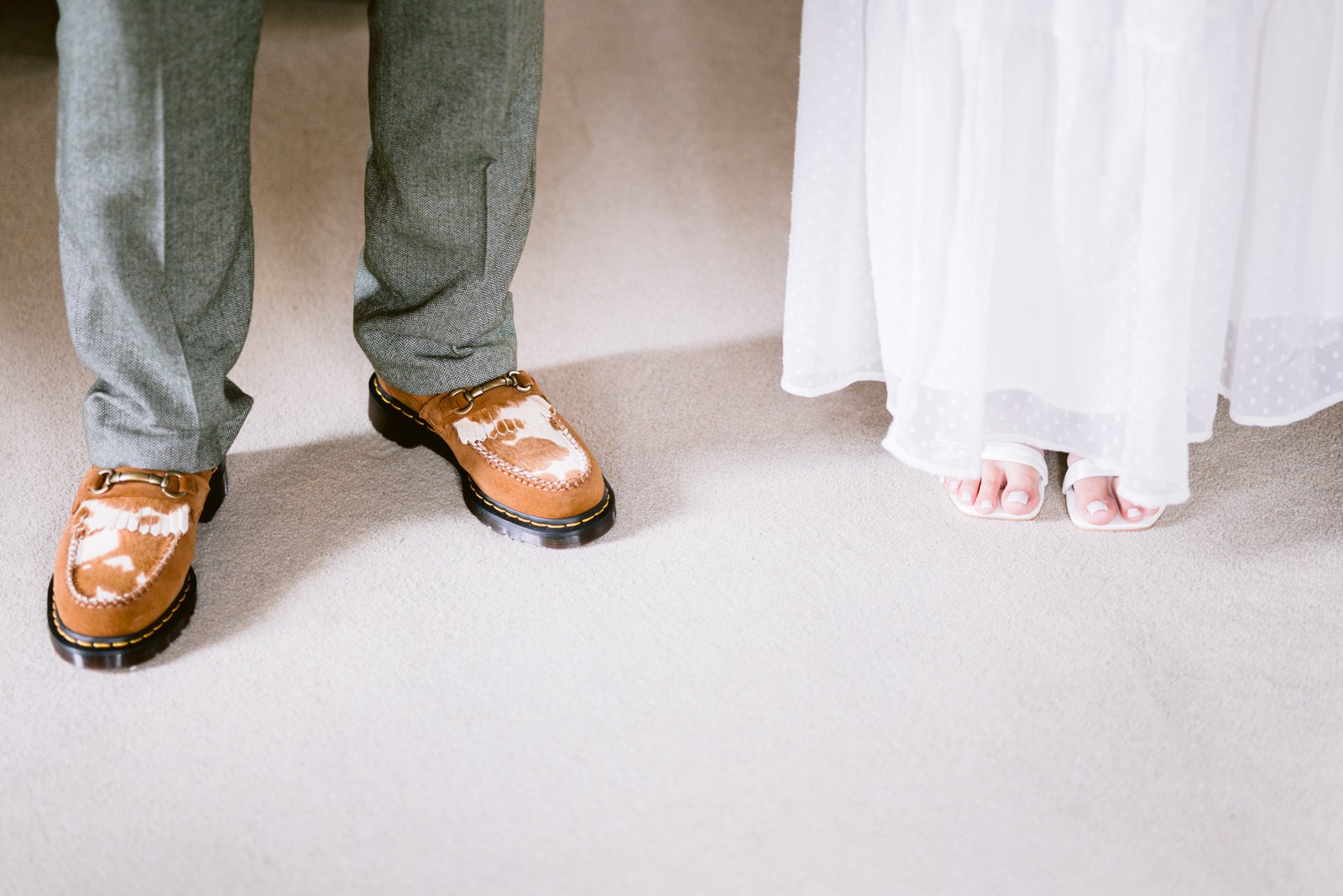 A person wearing gray pants with brown and white shoes stands next to another person in a white dress and white shoes, possibly attending a Carlisle Wedding.