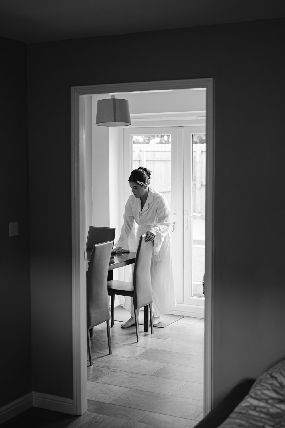 A relaxed person in a robe stands near a dining table with chairs in a room with patio doors, seen through a doorway, reminiscent of the tranquility before a Skitby Wedding.