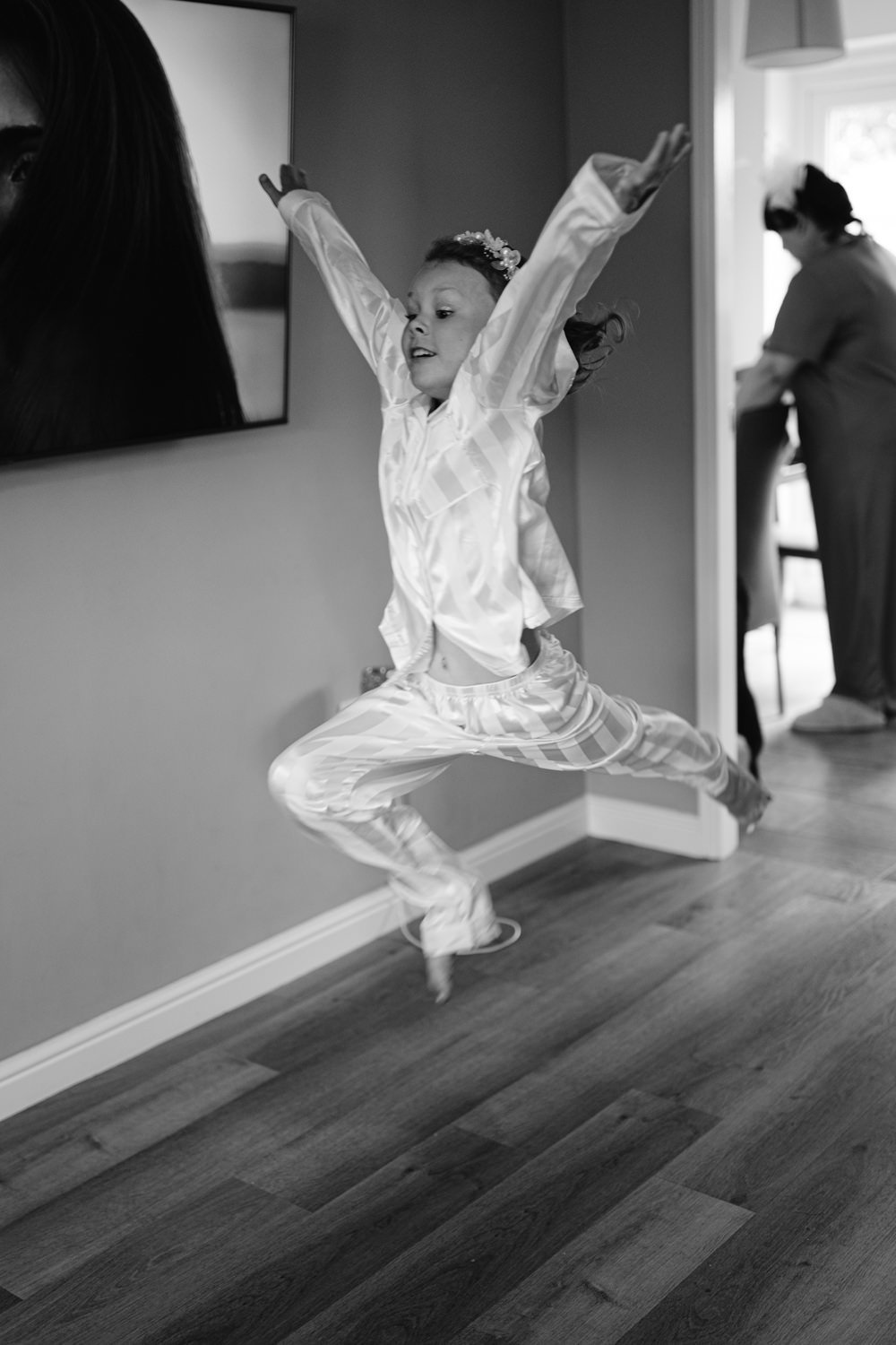 A young child, dressed in a shiny outfit, leaps into the air with arms outstretched at a relaxed wedding, while a person stands in a doorway in the background.
