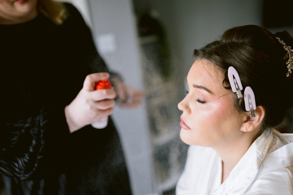 A woman with hair clips is having her hair styled by another person, who is spraying her hair with an aerosol spray. The woman has her eyes closed and appears relaxed, as if preparing for a fun Skitby Wedding.