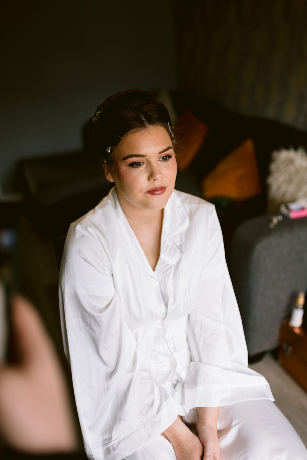 A person sits on a chair wearing a white robe, looking to the side with a neutral expression. Cushions and a couch adorn the background, adding to the relaxed wedding vibe.