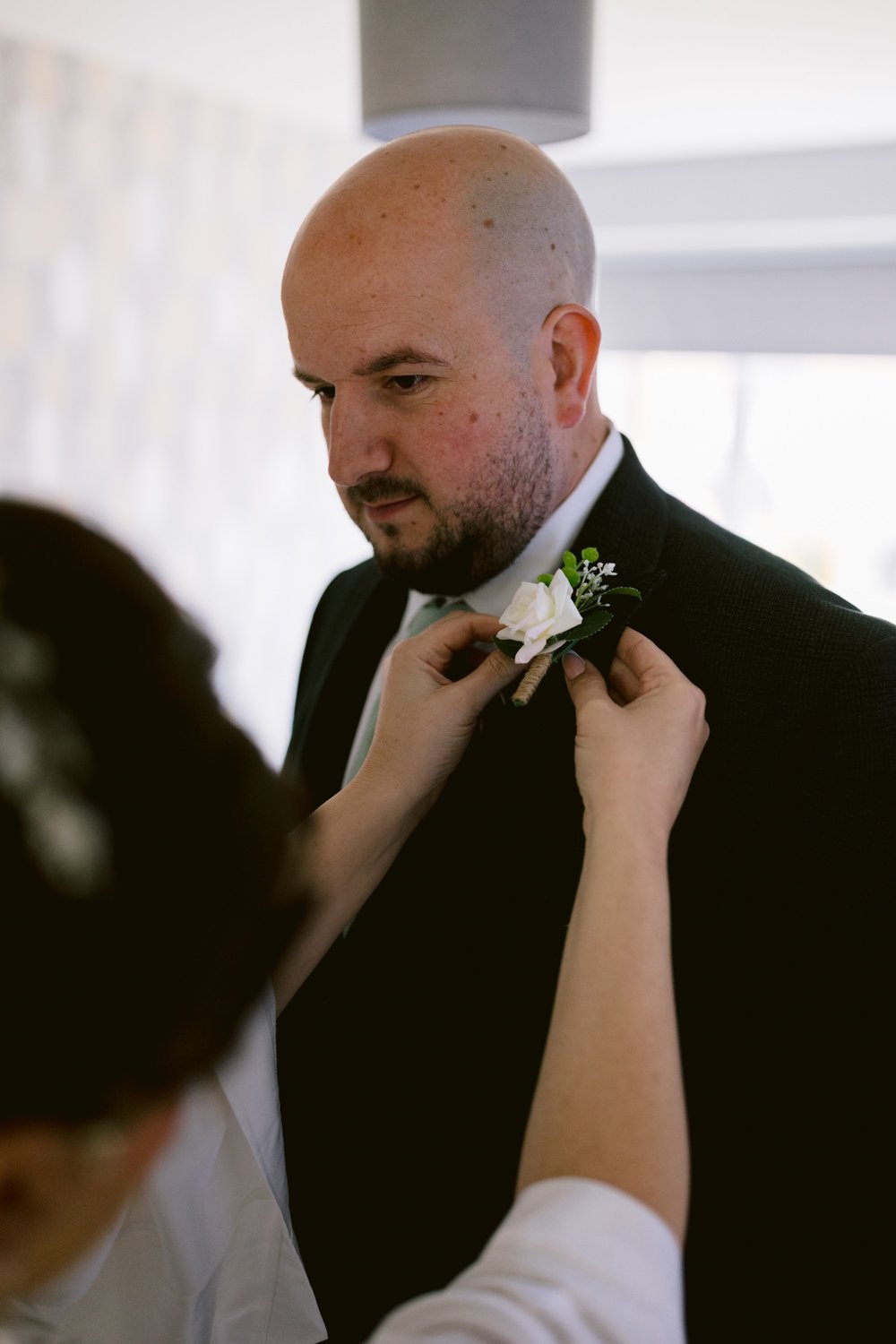 A person is pinning a boutonniere on a man in a suit inside a room at a relaxed wedding.