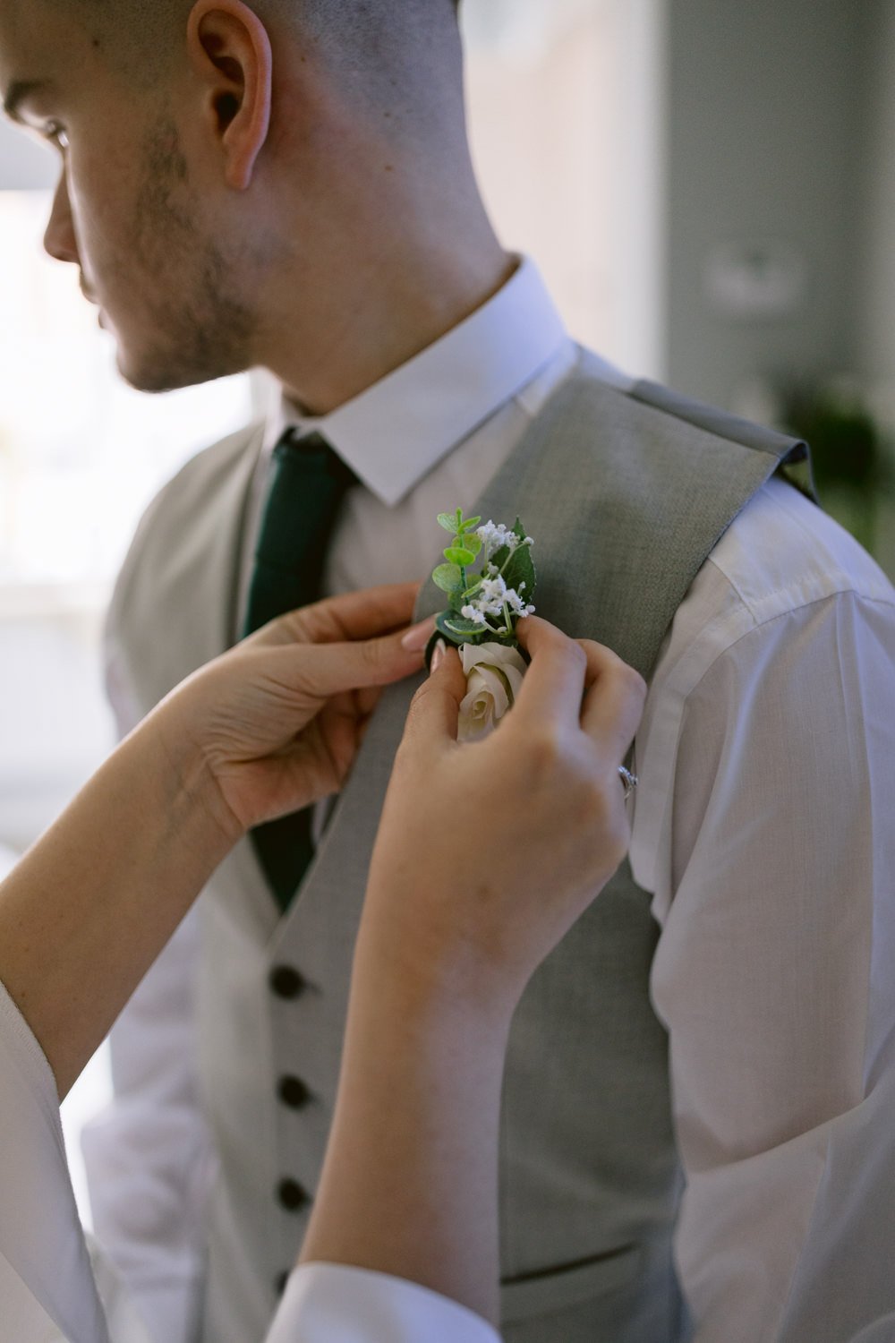 Person in a grey suit and white shirt with a boutonniere being pinned to their vest by another individual at a Skitby Wedding, creating a relaxed and fun atmosphere.