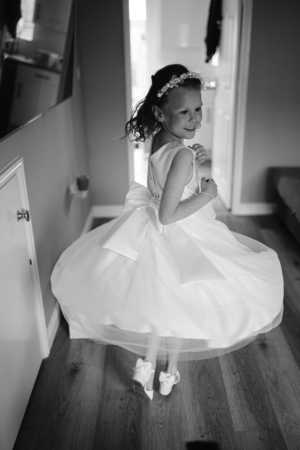 A young girl wearing a white dress and headband spins around joyfully in a room with wooden floors, capturing the essence of a fun wedding atmosphere.