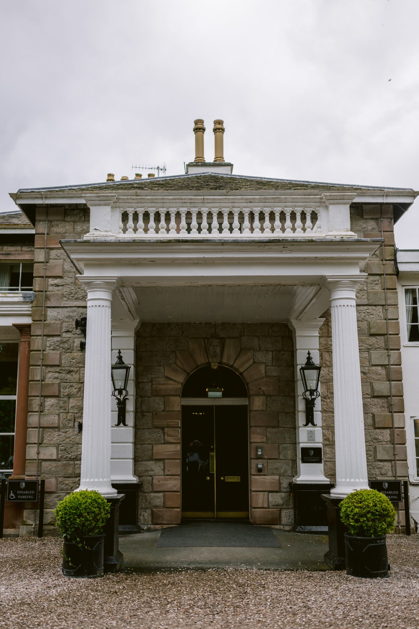 Front entrance of a historic stone building featuring tall white columns, lantern-style wall lights, and manicured bushes on either side of the doorway under a cloudy sky—perfect for an intimate Leeming House Micro Wedding.