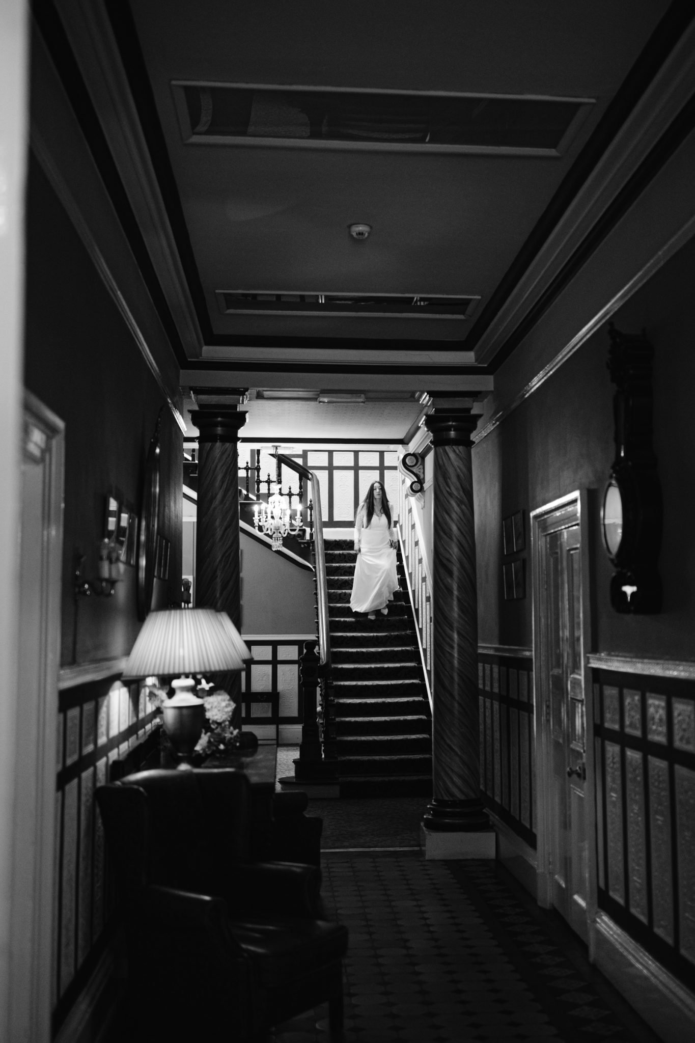 A person in a white dress ascends a staircase in a dimly lit, ornate hallway with pillars and a lamp on a table nearby, reminiscent of the elegance of Leeming House Micro Wedding. The scene is captured in black and white.
