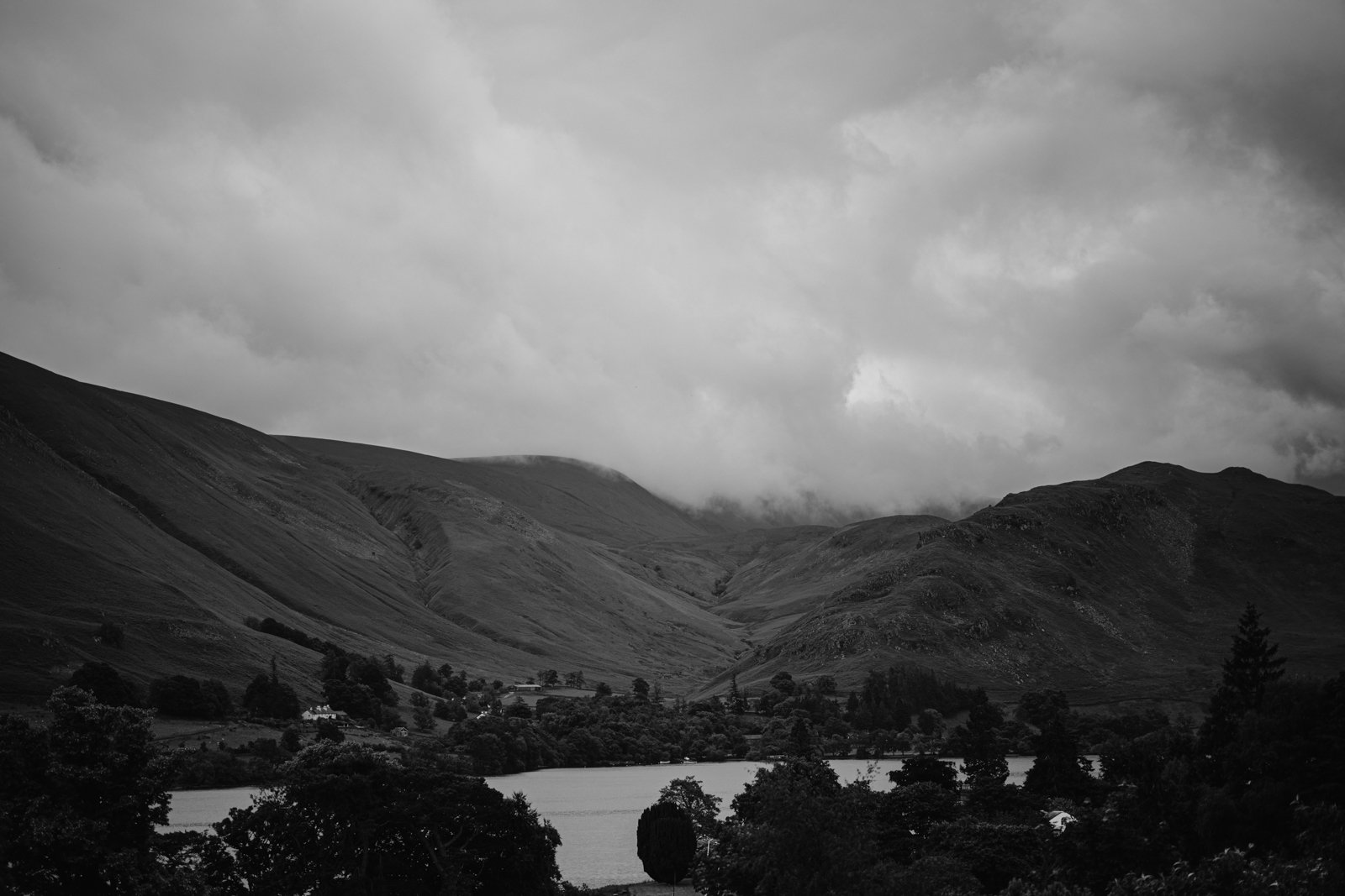 A black and white image of a landscape featuring hills and a valley under a cloudy sky, with a body of water and trees in the foreground, capturing the serene beauty perfect for an intimate Leeming House micro wedding.