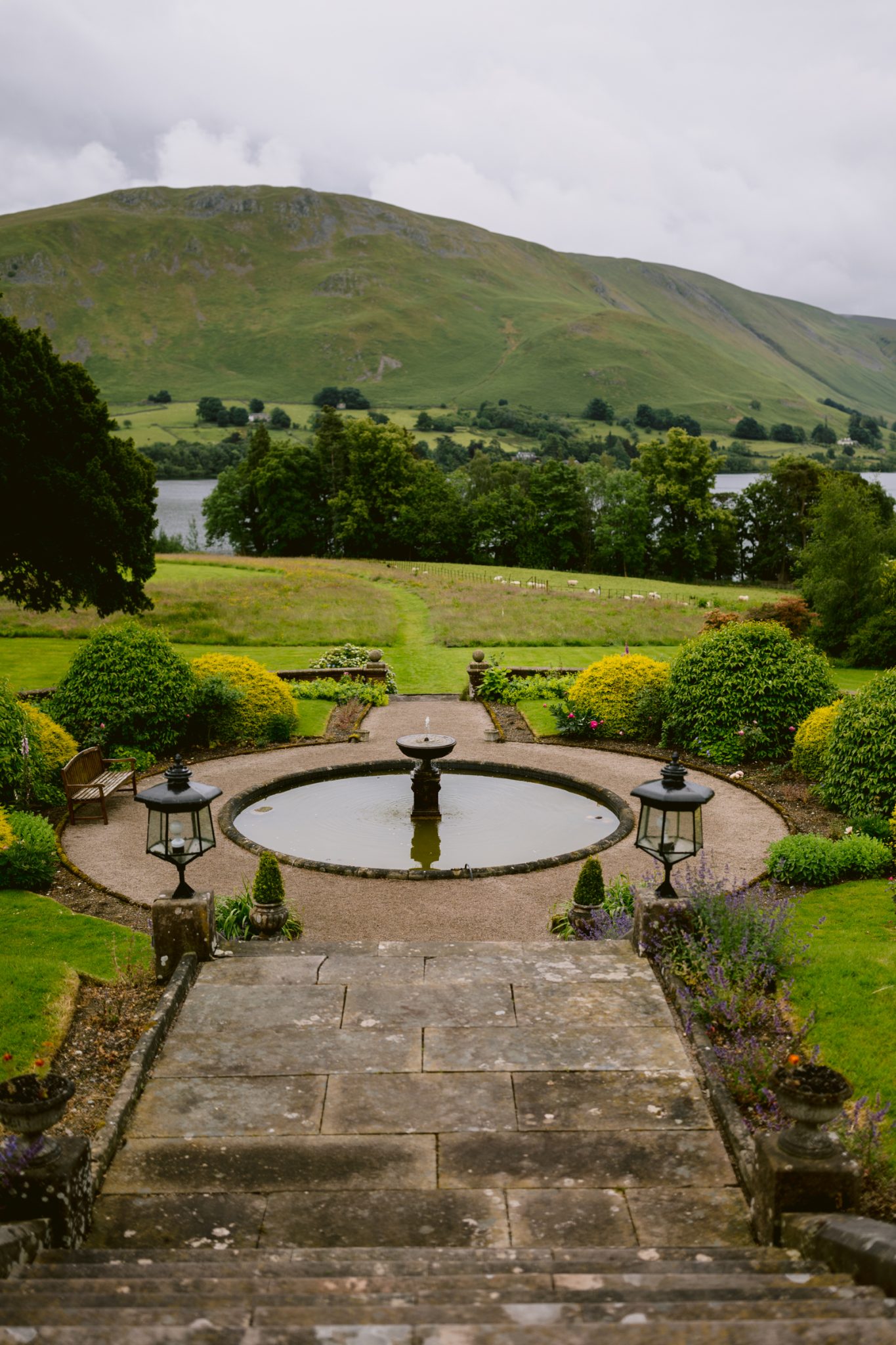 A circular fountain is centered in a landscaped garden with stone steps leading down to it; in the background, there's a grassy hill and a lake, making it an idyllic setting for a Leeming House micro wedding.