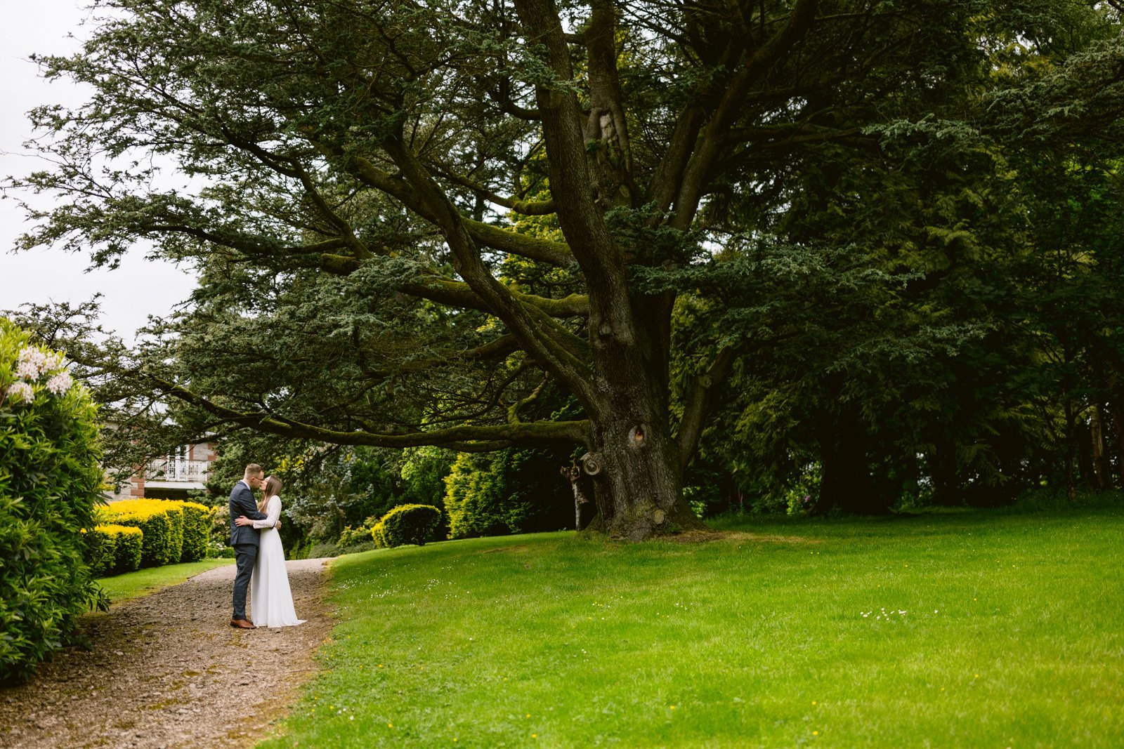 A couple dressed in wedding attire stands on a path under a large tree in a park-like setting, surrounded by greenery, capturing the intimate essence of a Leeming House Micro Wedding.