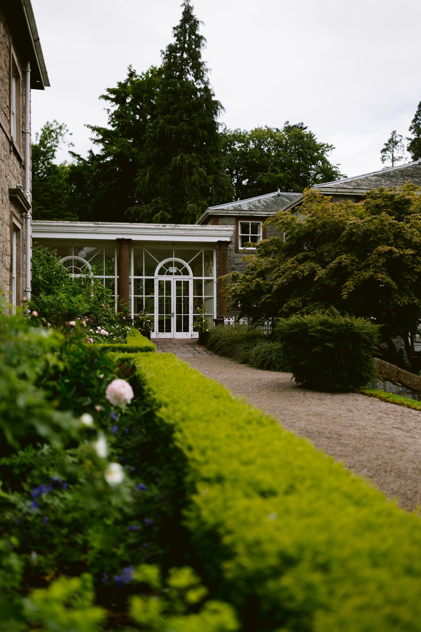 A glass-paned garden conservatory with white doors is flanked by greenery and connected to a stone building; the perfect setting for a Leeming House Micro Wedding. A picturesque gravel path leads to the entrance.