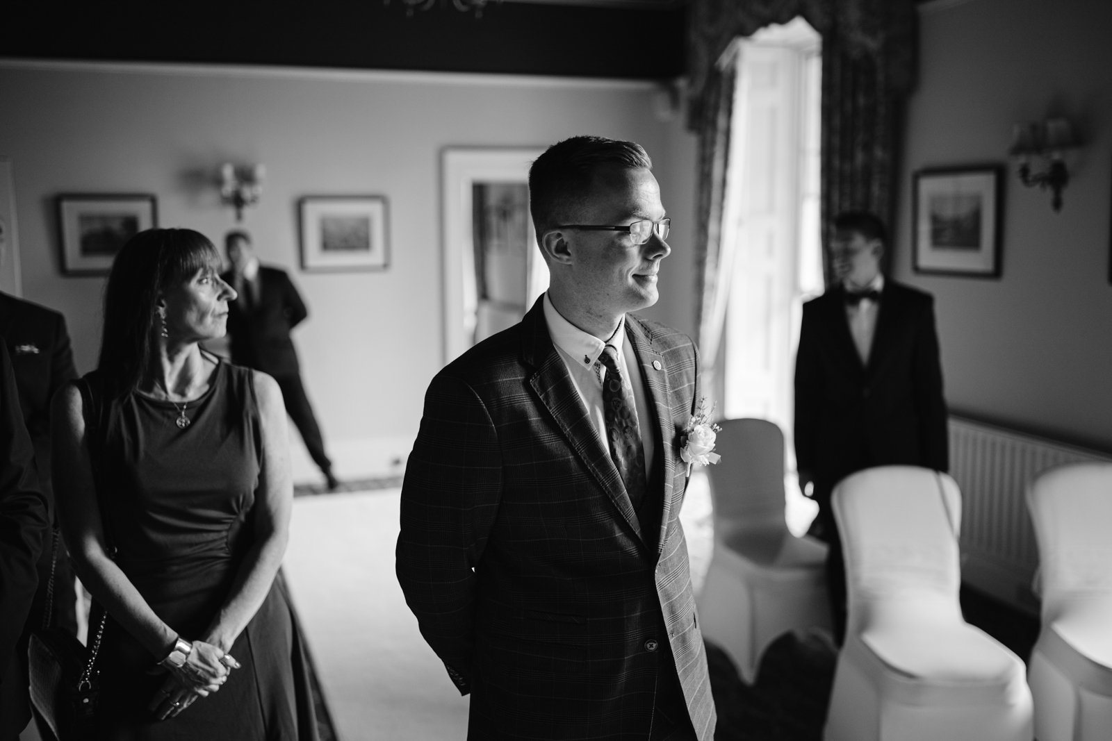 A man in a suit stands smiling in a room, accompanied by others. The setting appears to be formal, with empty chairs and framed pictures on the walls, reminiscent of an elegant Leeming House micro wedding.