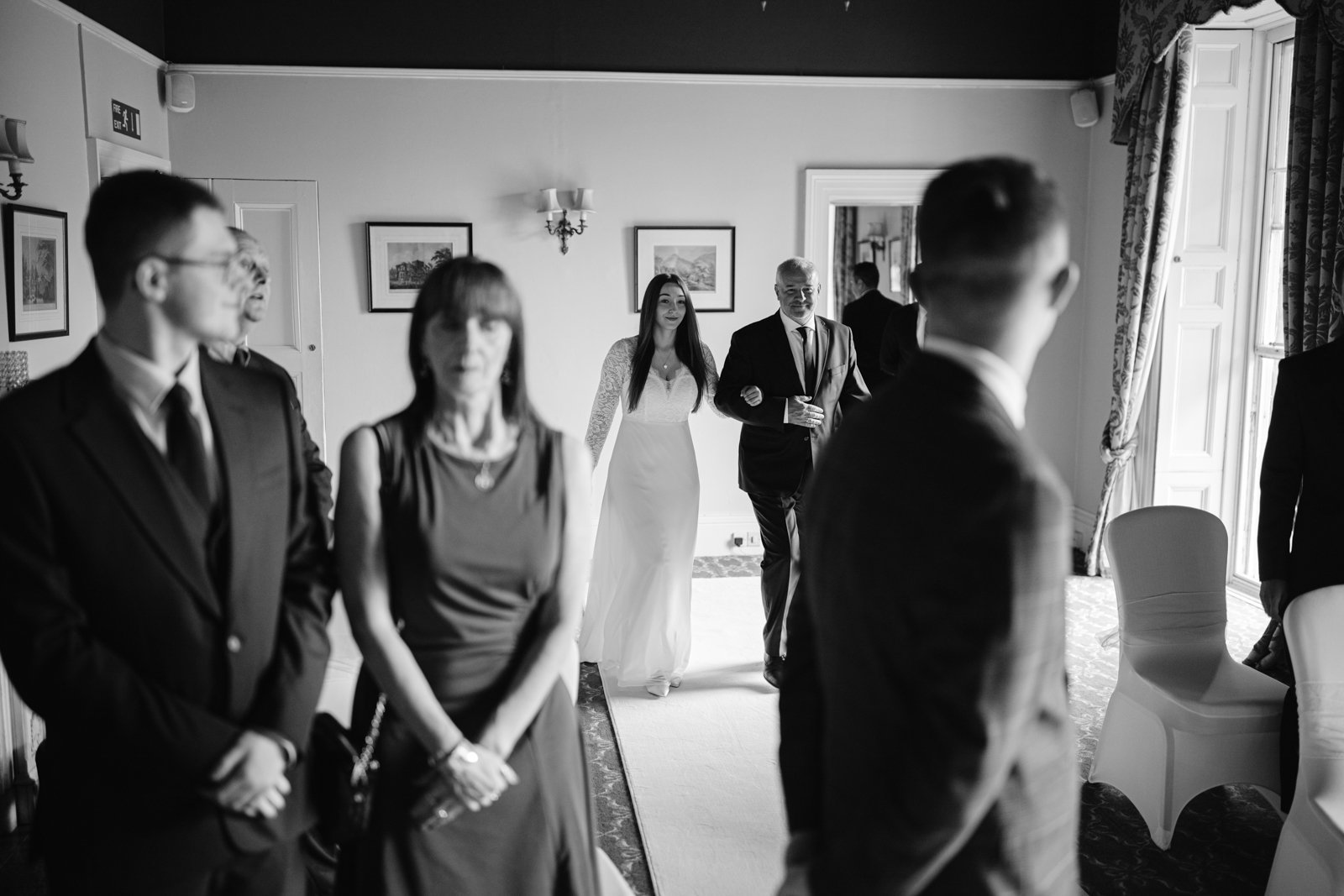 A bride walks down the aisle with an older man in a suit at a Leeming House Micro Wedding, while guests in formal attire are seated and standing on either side of a well-lit room decorated with framed pictures.