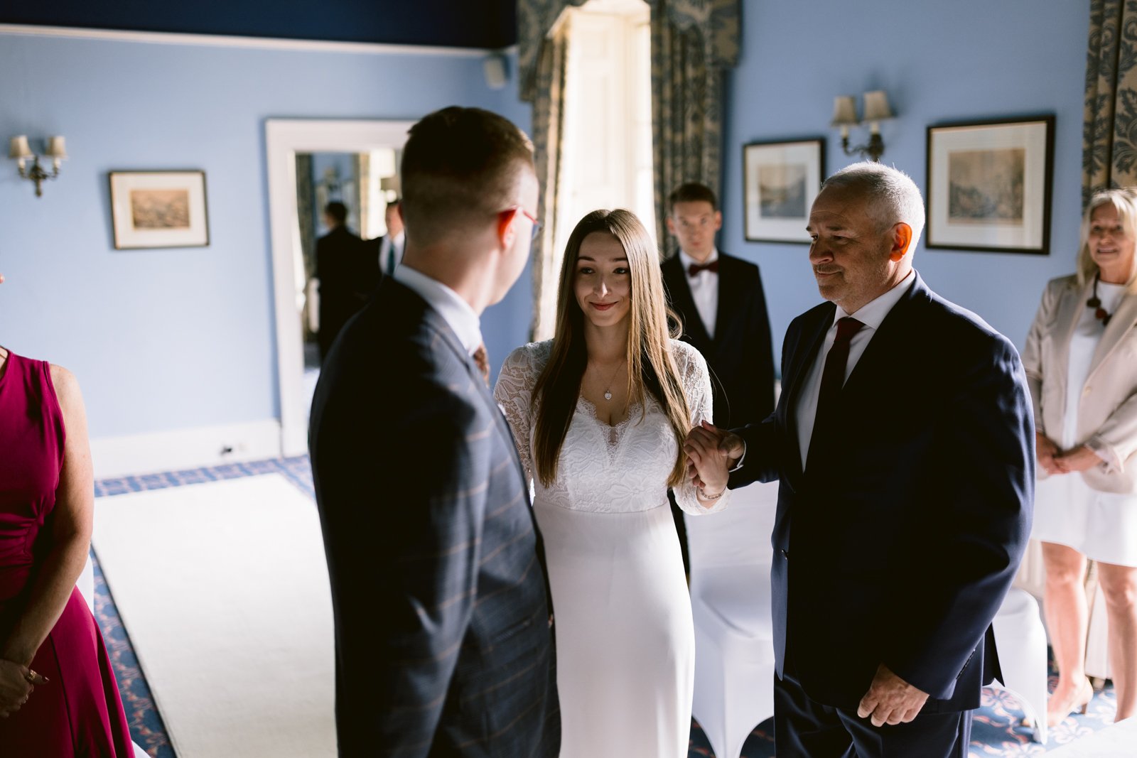 A bride in a white dress stands in the middle, with a groom in a suit on her right and an older man in a suit on her left. Other people are visible in the background inside a room with blue walls, capturing the intimate charm of their Leeming House Micro Wedding.