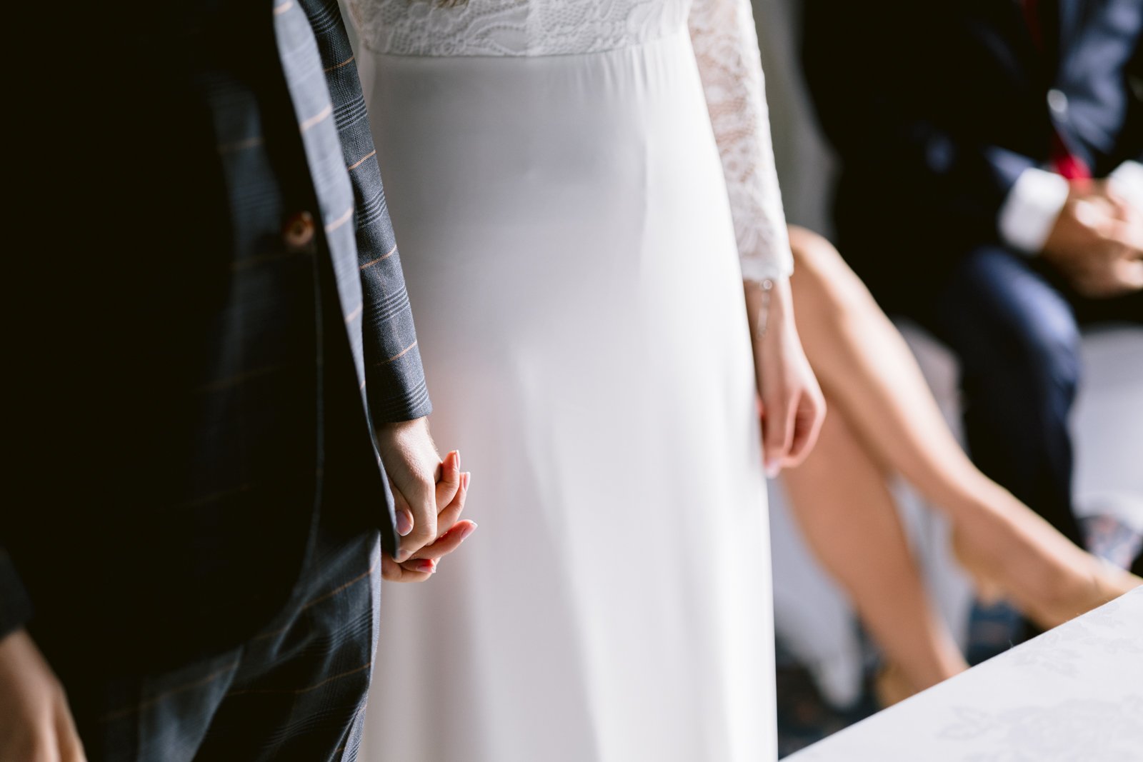 A couple holding hands, dressed in formal wedding attire, stands next to a seated person at their intimate Leeming House micro wedding.