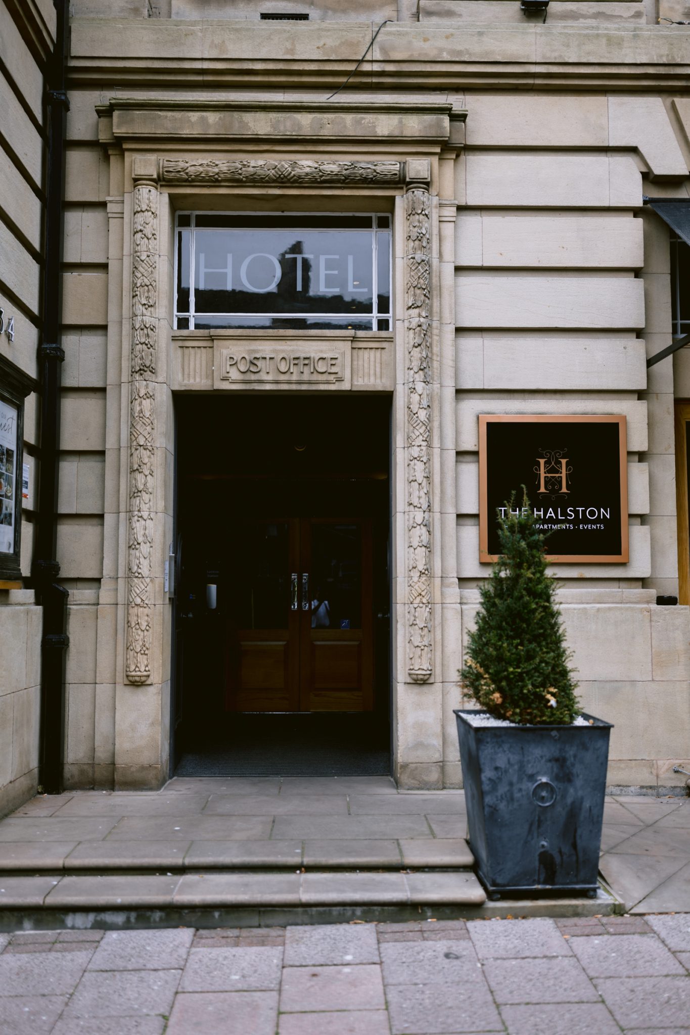 Entrance to a building with a sign above the door reading "HOTEL" and a partially visible sign to the right reading "THE HALSTON." A decorative potted plant is positioned at the front, adding charm to this castle town woodland setting perfect for a wedding.