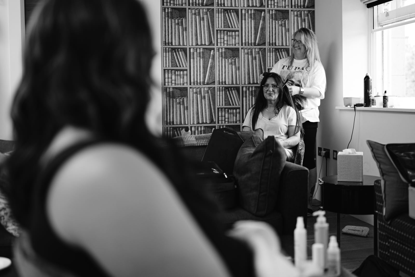 A woman sits on a couch getting her hair styled by another woman standing behind her, preparing for a woodland wedding. A person in the foreground is out of focus, facing the scene.