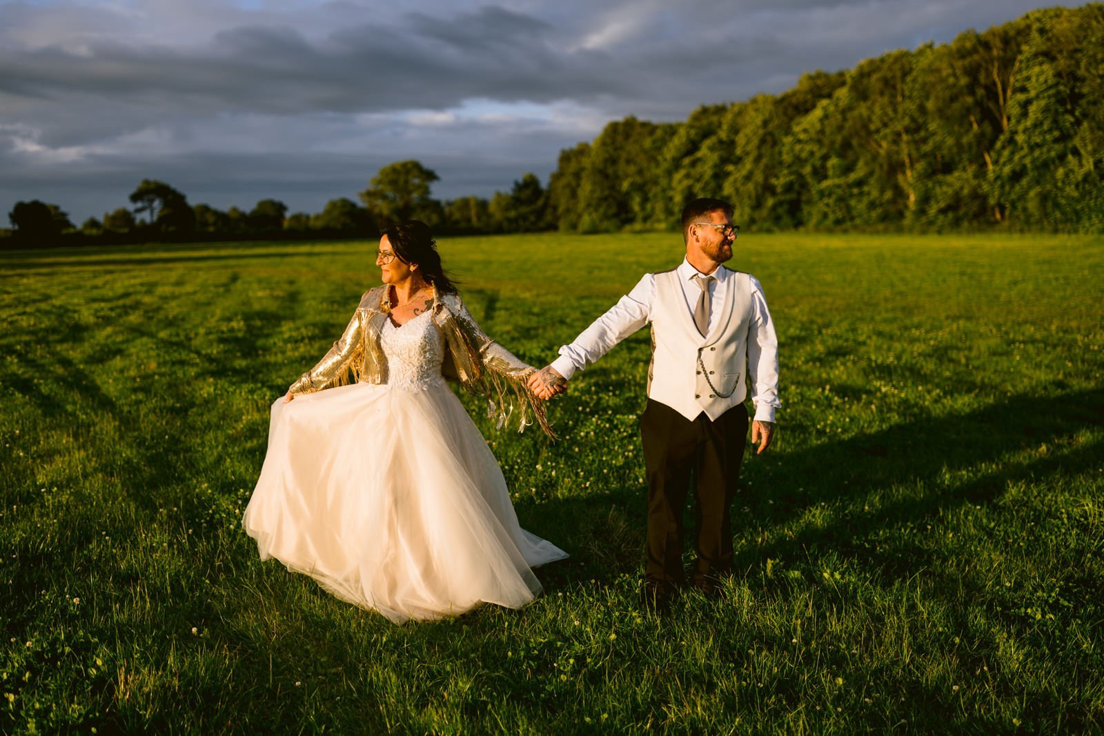 A couple in formal attire holds hands while standing on a grassy field, with the woman wearing a wedding dress and the man in a vest and tie. Trees of the woodland and a cloudy sky are visible in the background, evoking the timeless charm of a castle town wedding.