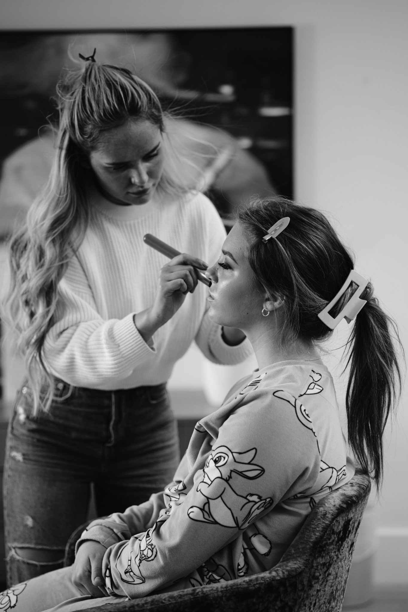 A woman applies makeup to the face of another woman who is seated. The seated woman, donned in a hair clip and a patterned top featuring cartoon characters, appears as if she's preparing for a whimsical woodland wedding.