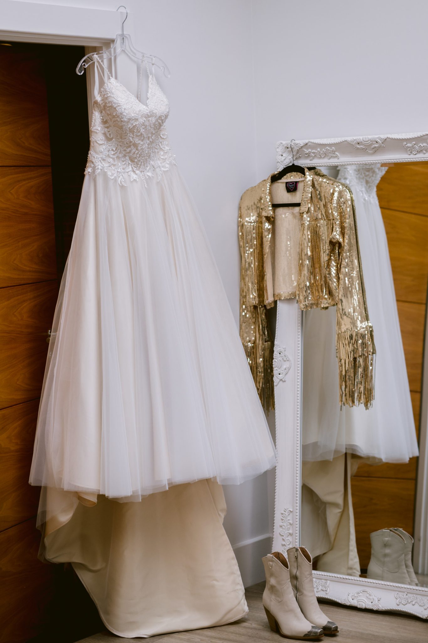 A white wedding dress and a gold sequined jacket hang next to a full-length mirror, with a pair of beige boots placed on the floor below them, evoking a woodland wedding in a charming castle town.
