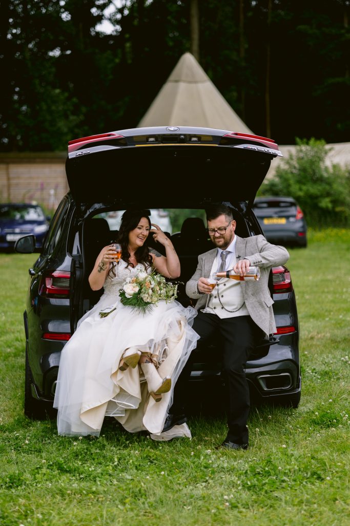 A couple in wedding attire sit on the open tailgate of a black SUV, smiling and holding drinks, against the picturesque backdrop of a woodland wedding near a charming castle town.
