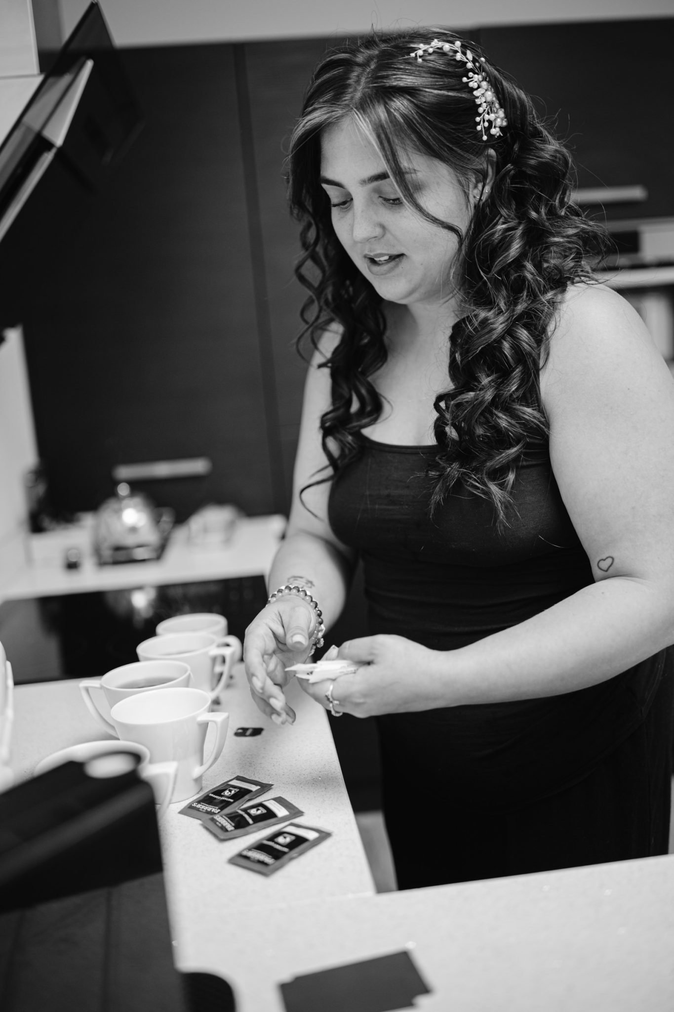 A woman in a kitchen prepares tea, holding a tea bag and standing in front of several cups. She has long, wavy hair and is wearing a sleeveless dress, reminiscent of a serene woodland wedding.