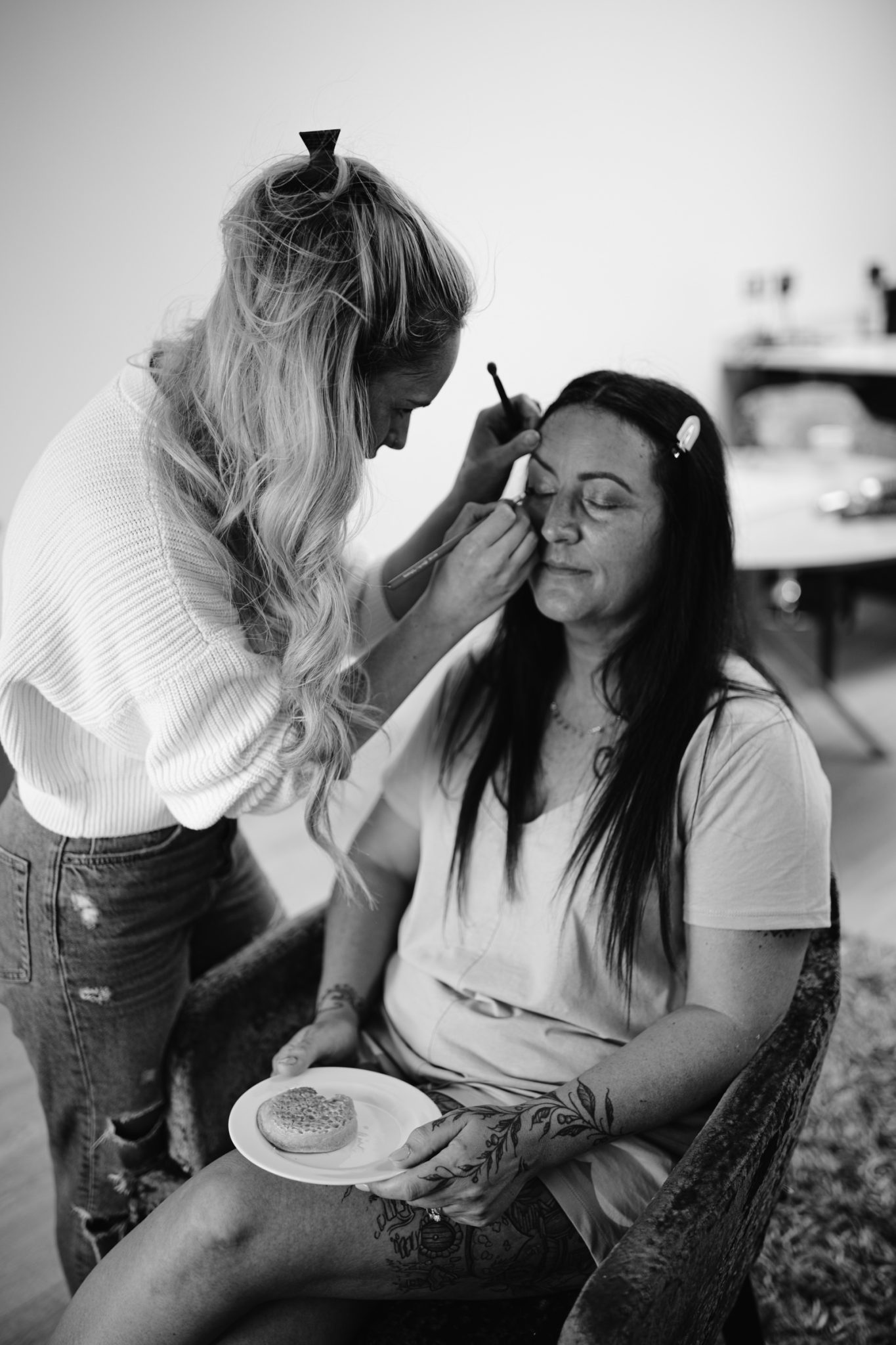 A woman is having makeup applied by another woman. She is seated in a cozy room, holding a plate with a partially eaten sandwich, while the makeup artist deftly works on her face. The atmosphere feels enchanting, as if preparing for a grand woodland wedding in an ancient castle town.
