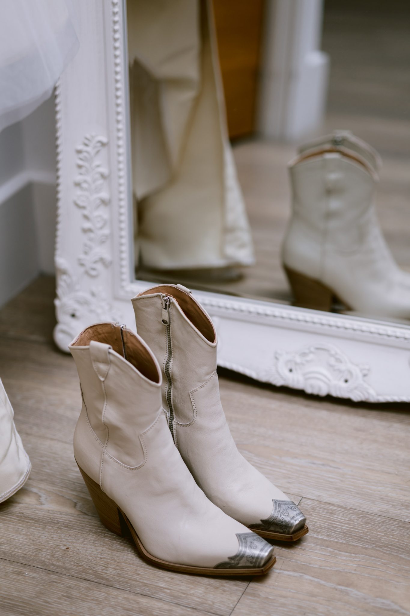 A pair of white cowboy boots with metallic toe caps is placed on a wooden floor in front of an ornate white mirror, reflecting the boots like a scene from a rustic woodland wedding.