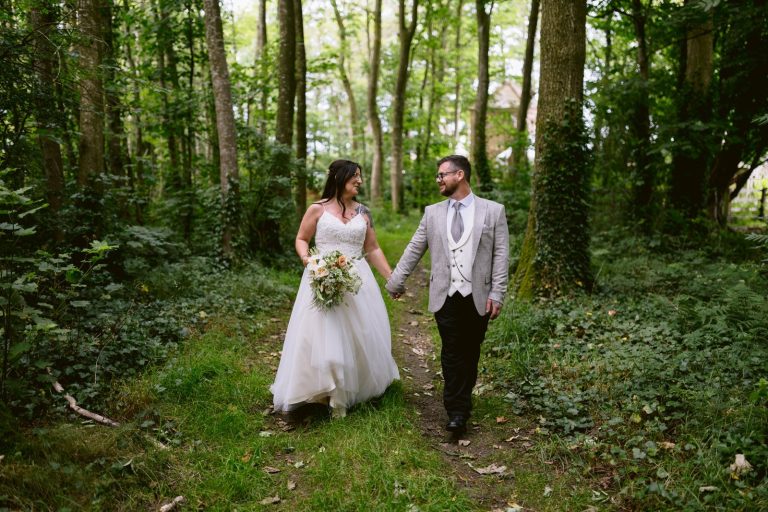 A couple in wedding attire holds hands while walking through a castle town's woodland path. The bride wears a white dress and carries a bouquet, and the groom dons a gray suit with a tie.