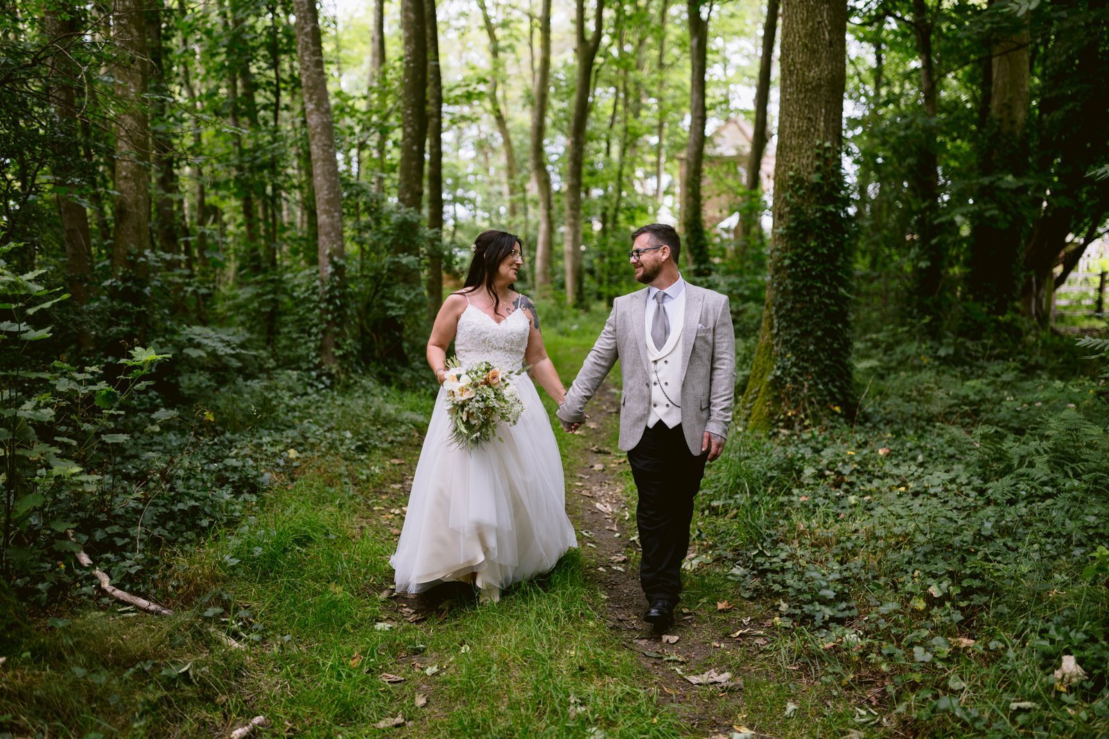 A couple in wedding attire holds hands while walking through a castle town's woodland path. The bride wears a white dress and carries a bouquet, and the groom dons a gray suit with a tie.