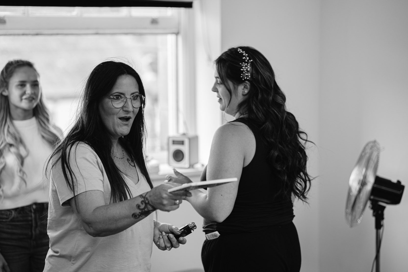 A woman holding a clipboard speaks emphatically to another woman with long hair, who faces away. A third woman smiles in the background. The room, reminiscent of a quaint woodland cabin, features a small fan and a camera.