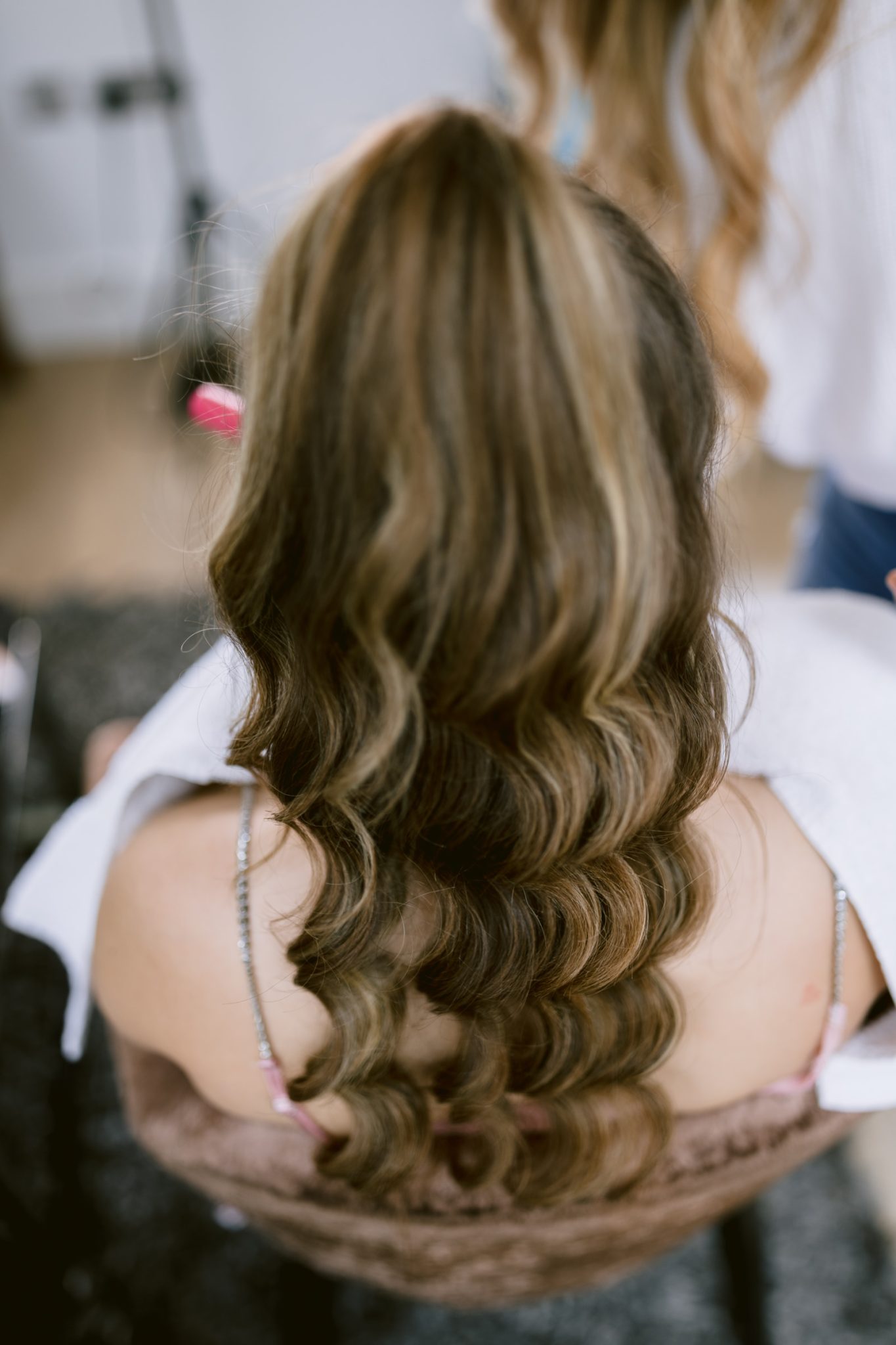 A woman with long wavy hair, highlighted with streaks of blond, is seen from behind while getting her hair styled for a woodland wedding.