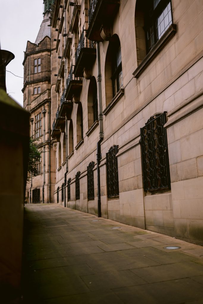 Stone building with arched windows and decorative ironwork stands regally, reminiscent of the Sheffield Registry Office where intimate weddings come alive, set along a narrow, empty sidewalk under a cloudy sky.