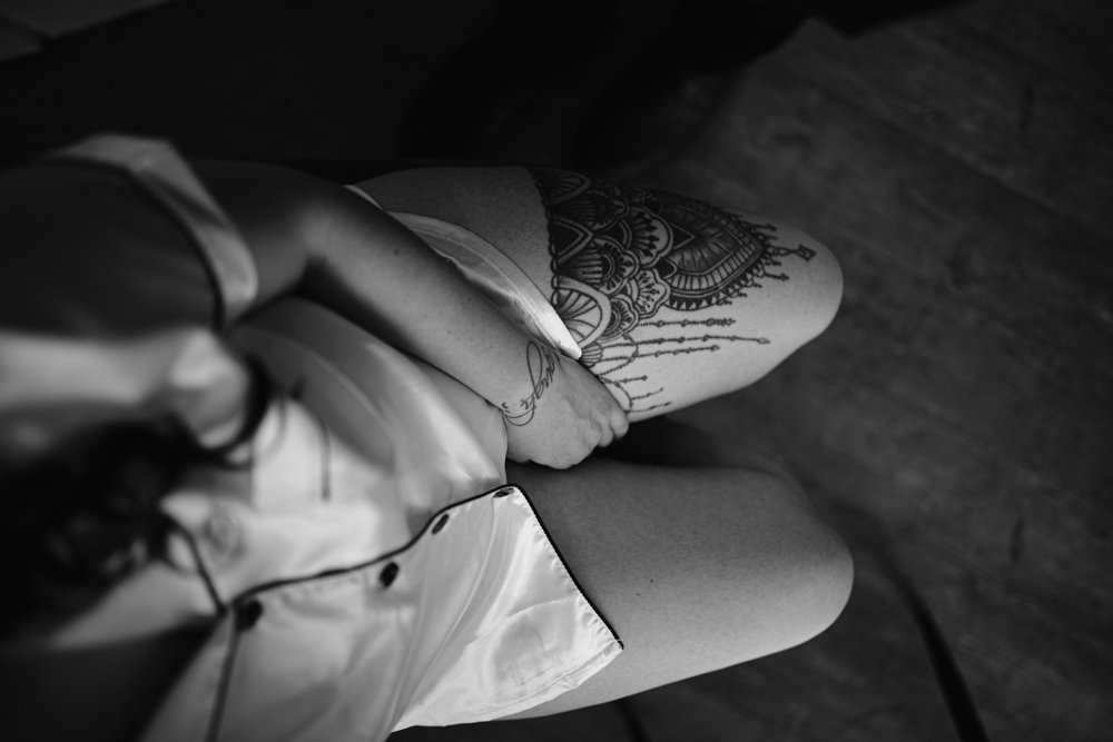 Black and white image of a person sitting with one leg crossed over the other, showing an intricate tattoo on the upper thigh. They are wearing a short-sleeved shirt and shorts.