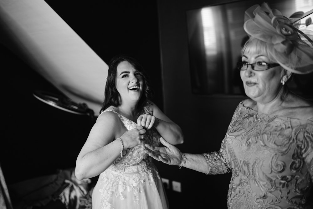 Bride in a white dress laughing, standing with a woman in a floral hat and glasses, inside a dimly lit room.