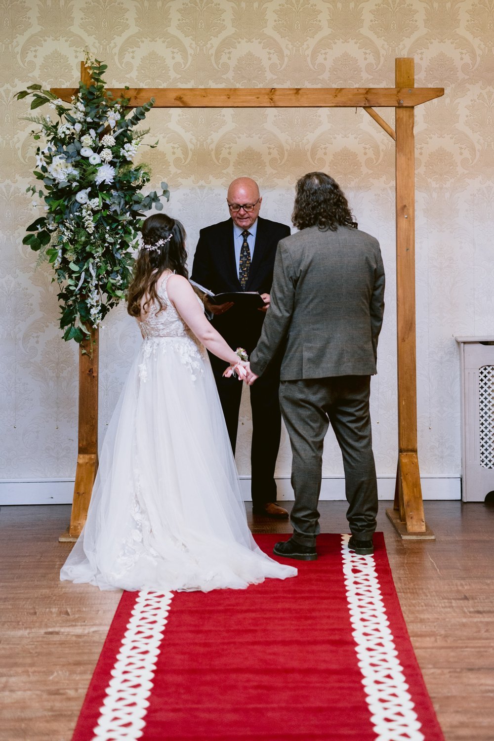 A couple stands holding hands under a wooden arch adorned with flowers, facing an officiant. They are on a red carpet, preparing to exchange vows in a wedding ceremony.