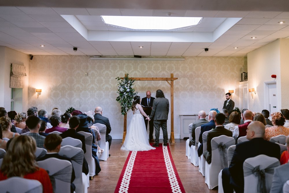 A bride and groom stand at the altar in a decorated room, facing an officiant. Guests are seated on either side of an aisle with red carpet.