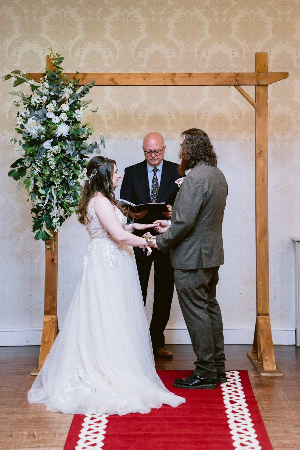 A couple stands under a wooden arch during a wedding ceremony. The officiant, holding a book, faces them. The bride wears a white gown and the groom is in a dark suit.