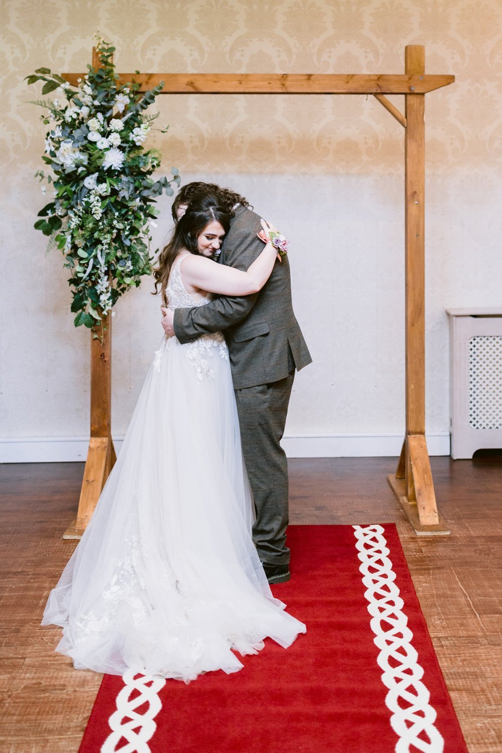 Bride and groom embracing under a wooden arch adorned with flowers, standing on a red carpet with white patterns.
