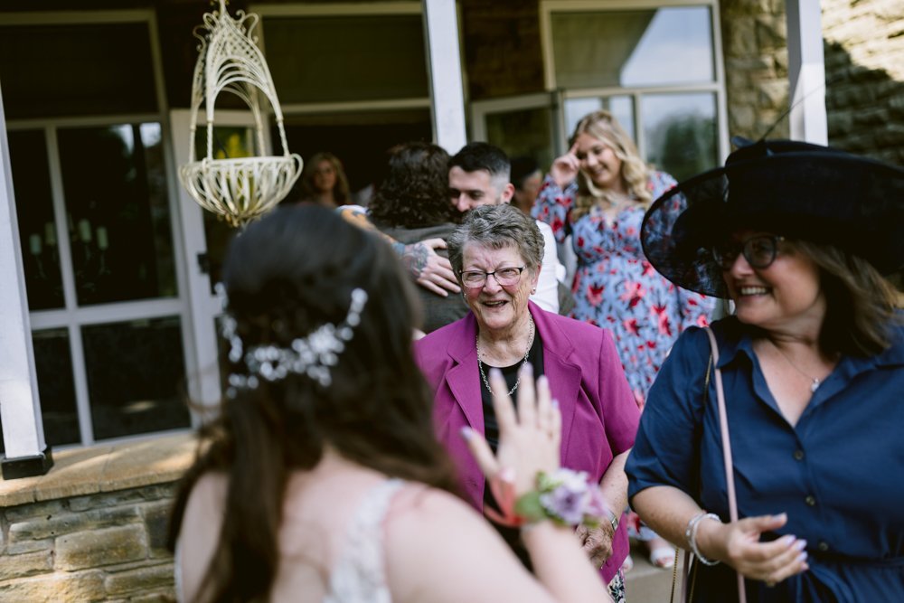 Group of people outdoors, some hugging. A woman in a pink jacket smiles, another in a black hat looks on.