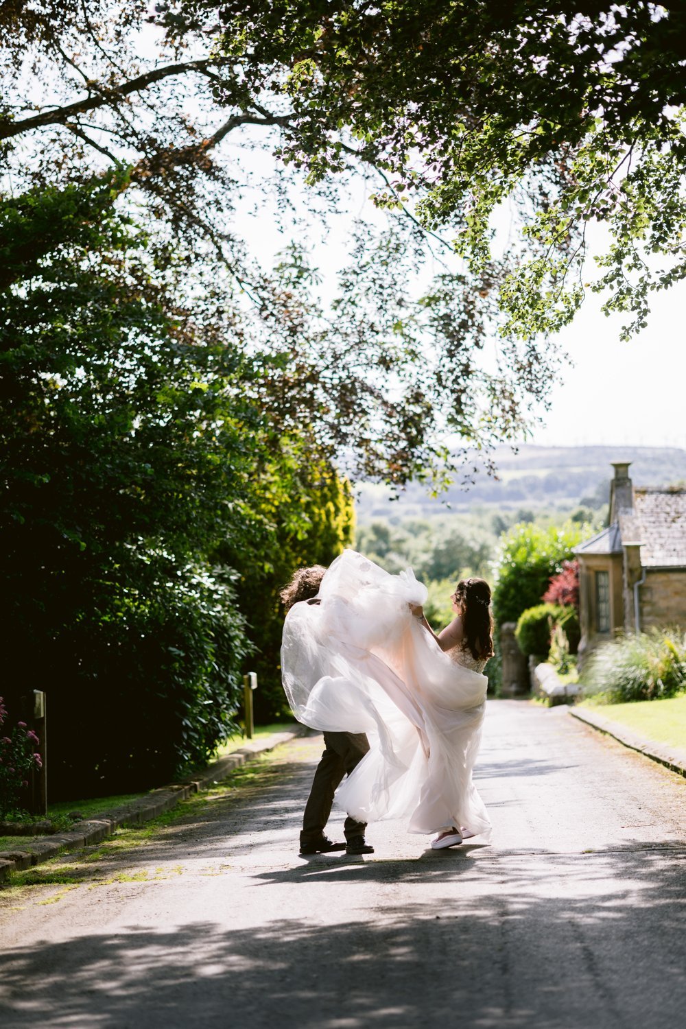 A couple in wedding attire playfully walking on a sunlit path, surrounded by greenery and a stone building in the background.