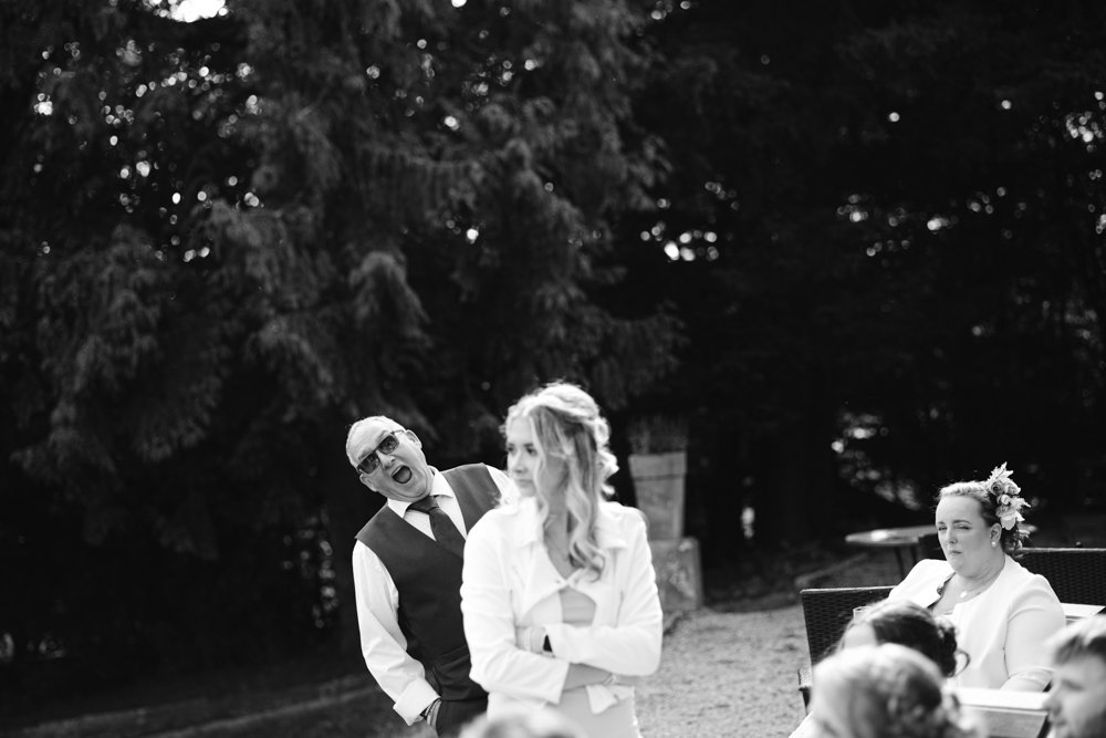 A man in a vest and tie makes a surprised face behind a woman in a dress, while another woman sits nearby in an outdoor setting.
