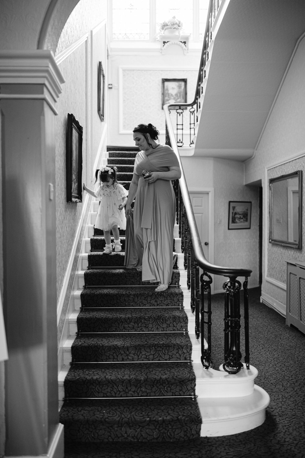 Woman and young girl in a dress walking down a carpeted staircase in a dimly lit interior with framed artwork on the walls.
